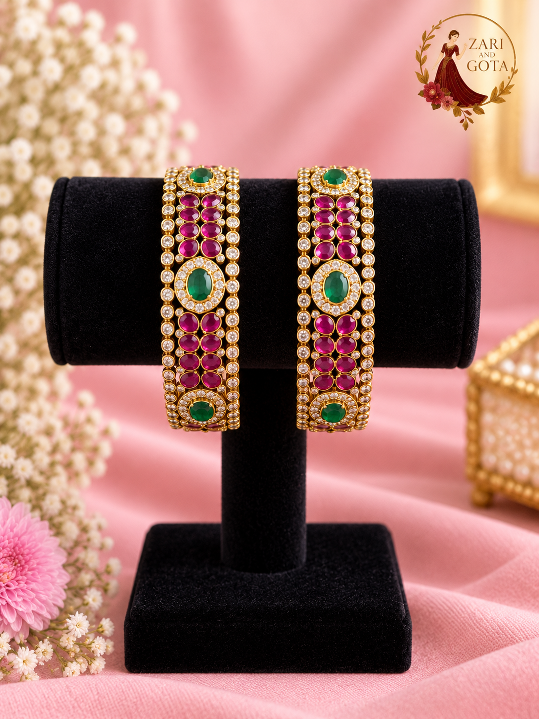 Pair of wide gold-tone bangles with rows of ruby-pink stones, oval emerald green centre stones and American diamond borders on a black velvet stand, Zari and Gota UK